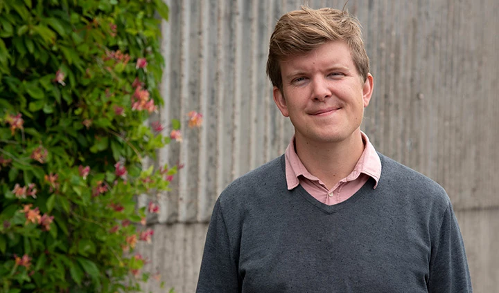 Man i grå tröja, står framför en mur, intill en klätterväxt